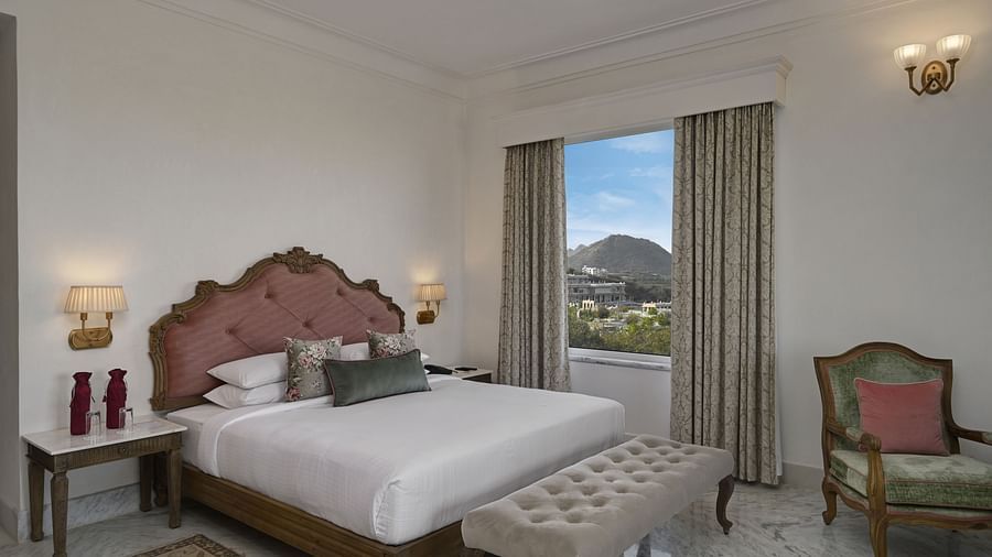 A well-lit Heritage Chamber Suite at Khas Mahal by Fateh Collection, Udaipur with a large bed, a tufted bench at the foot of the bed, a chair, a nightstand with a lamp, and a window offering a view of an outdoor landscape.