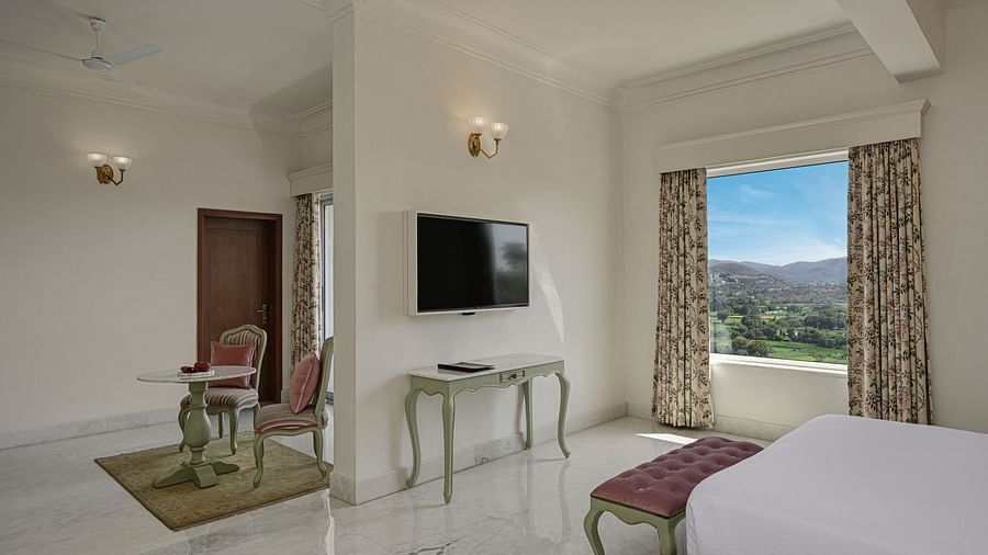 Close up of a  well-lit Heritage Chamber Suite at Khas Mahal by Fateh Collection, Udaipur with a large bed, a tufted bench at the foot of the bed, a chair, a nightstand with a lamp, and a window offering a view of an outdoor landscape.