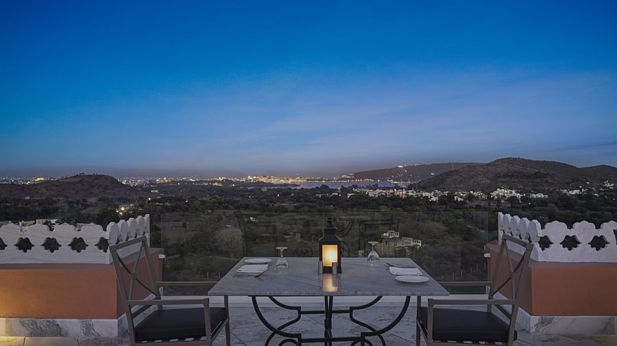 An outdoor table and chairs on a balcony or patio, with a view of a landscape in the distance at Khas Mahal by Fateh Collection, Udaipur. 
