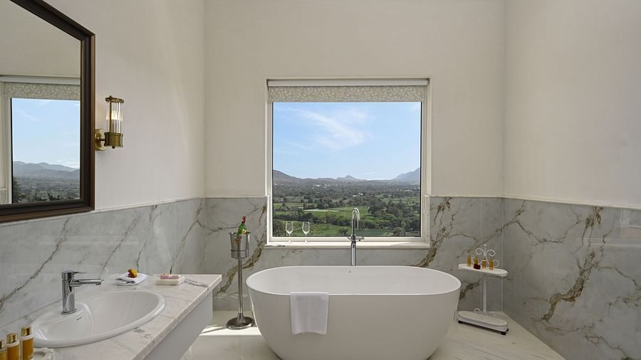 A well-lit, modern bathroom featuring a freestanding bathtub, a marble-topped vanity with a sink, and a window offering an outdoor view at Heritage Chamber Suite at Khas Mahal by Fateh Collection, Udaipur.