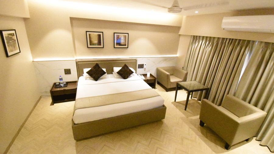 An Executive Room at Kings International Juhu featuring a bed, 2 sofas and a centre table, an A/C unit, and paintings on the walls.