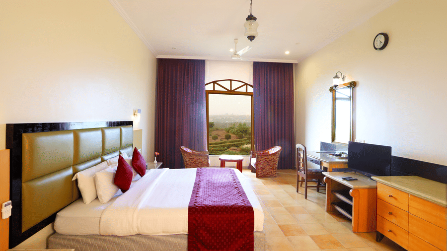 A bright hotel bedroom with a big bed, a desk and a TV, and 2 chairs by an arched window overlooking the greenery outside | Kohinoor Samudra Beach Resort, Ratnagiri