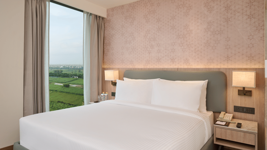 A bedroom at KPG Sarovar Portico, Raj Nagar Extension, Ghaziabad featuring a king size bed, side tables with lamps, a bedside telephone, and a window with curtains.