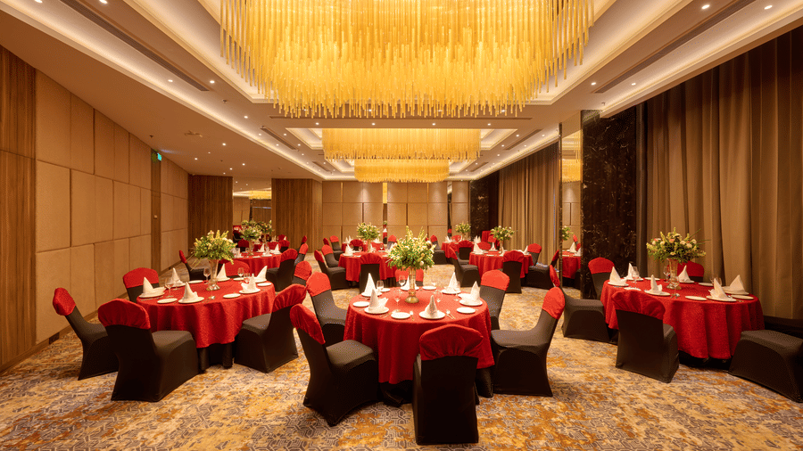 A large banquet hall at KPG Sarovar Portico, Raj Nagar Extension, Ghaziabad featuring round dining tables, chairs, chandeliers, and carpeted flooring.