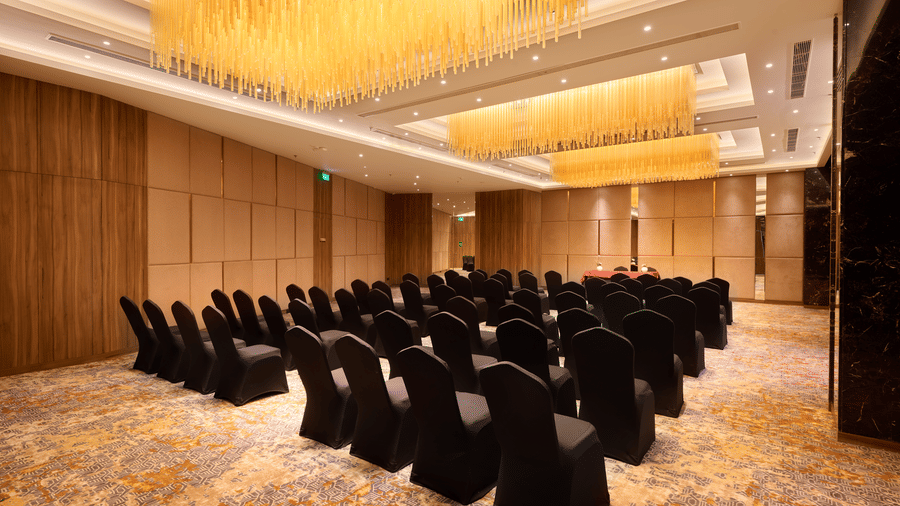 A conference hall setup at KPG Sarovar Portico, Raj Nagar Extension, Ghaziabad featuring rows of chairs, chandeliers, carpeted flooring, and a head table.