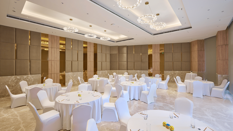 An event hall at KPG Sarovar Portico, Raj Nagar Extension, Ghaziabad featuring round tables, covered chairs, ceiling light fixtures, and tiled flooring.