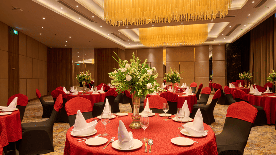 A banquet hall view at KPG Sarovar Portico, Raj Nagar Extension, Ghaziabad featuring round dining tables, chairs, flower arrangements, and chandeliers.