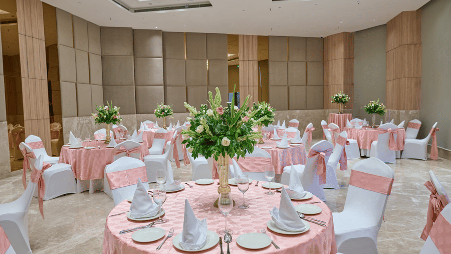 An event hall setup at KPG Sarovar Portico, Raj Nagar Extension, Ghaziabad featuring round dining tables, chairs with bows, flower vases, and wall panels.