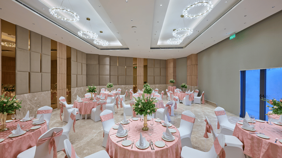 An event hall view at KPG Sarovar Portico, Raj Nagar Extension, Ghaziabad featuring round dining tables, chairs with bows, ceiling lights, and side tables.