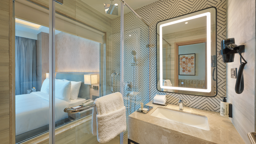 A bathroom at KPG Sarovar Portico, Raj Nagar Extension, Ghaziabad featuring a washbasin with countertop, an illuminated mirror, a glass shower partition, and a view into the bedroom.