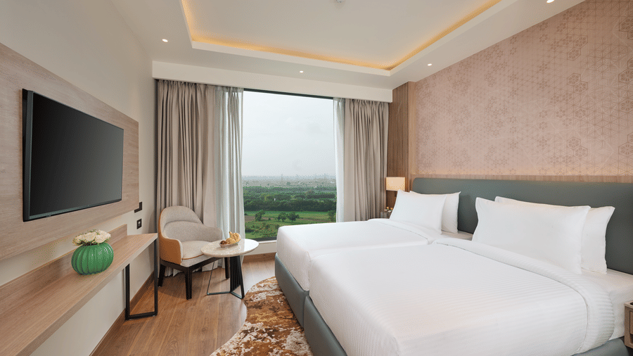 A bedroom at KPG Sarovar Portico, Raj Nagar Extension, Ghaziabad featuring twin beds, a wall mounted television, an armchair with a side table, and a large window with curtains.