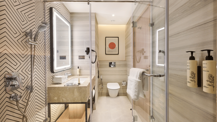 A bathroom at KPG Sarovar Portico, Raj Nagar Extension, Ghaziabad featuring a vanity sink, a illuminated mirror, a glass shower cubicle, and a wall mounted toilet.