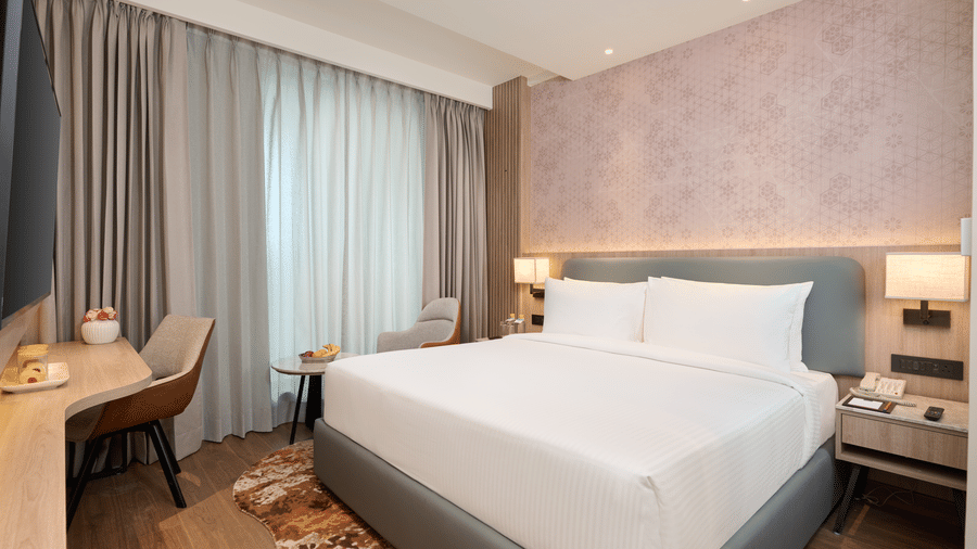 A bedroom at KPG Sarovar Portico, Raj Nagar Extension, Ghaziabad featuring a king size bed, a study desk with chair, a bedside table, and window curtains.