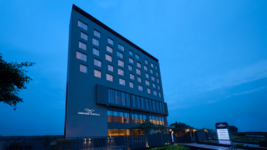 A hotel building exterior at KPG Sarovar Portico, Raj Nagar Extension, Ghaziabad featuring lighted windows, a front entrance, a sign board, and surrounding greenery.