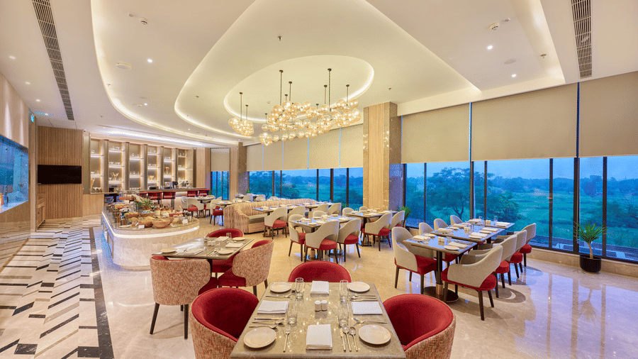 A dining area at KPG Sarovar Portico, Raj Nagar Extension, Ghaziabad featuring dining tables, chairs, a ceiling light, and large windows.