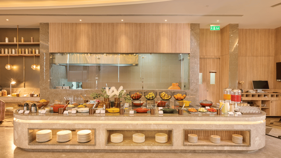 A buffet counter at KPG Sarovar Portico, Raj Nagar Extension, Ghaziabad featuring food dishes, serving bowls, juice dispensers, and under-counter plate storage.