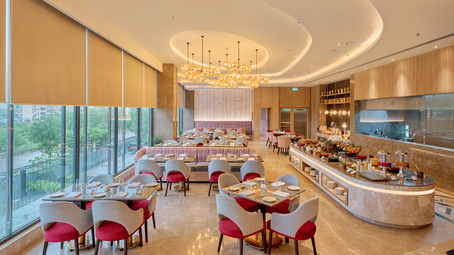 A restaurant interior at KPG Sarovar Portico, Raj Nagar Extension, Ghaziabad featuring dining tables, padded chairs, a long buffet counter, and window blinds.