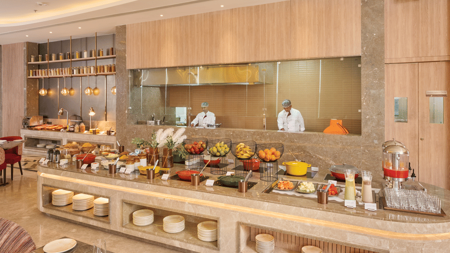 A live cooking counter at KPG Sarovar Portico, Raj Nagar Extension, Ghaziabad featuring 2 chefs behind a glass partition, food containers, plates, and juice dispensers.