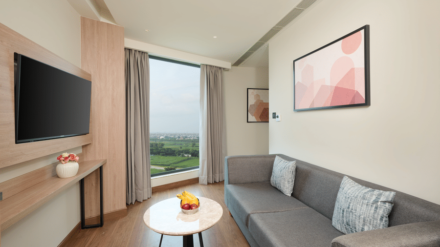A living room area at KPG Sarovar Portico, Raj Nagar Extension, Ghaziabad featuring a sofa, a coffee table, a wall mounted television, and wall artwork.