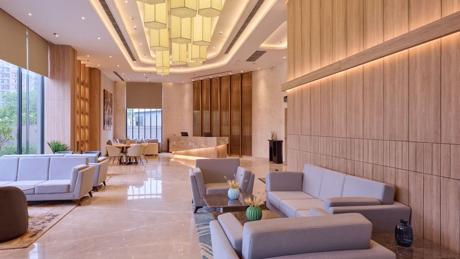 A hotel lobby at KPG Sarovar Portico, Raj Nagar Extension, Ghaziabad featuring sofa sets, coffee tables, ceiling lights, and wooden wall panelling. 
