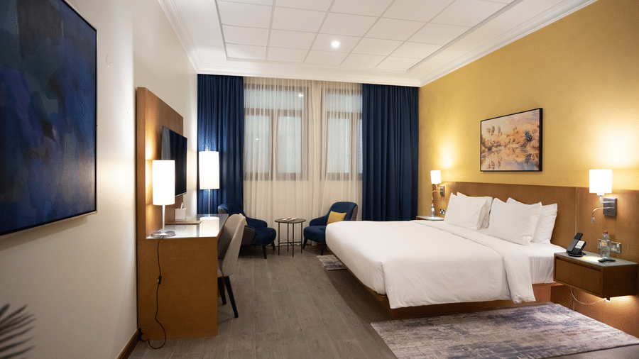 Wide view of a hotel-style room. Bed, yellow accent wall, windows with curtains, and a desk/TV unit with a blue abstract painting nearby.