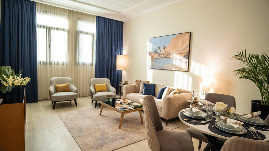 Bright living and dining room. Neutral sofa, two grey chairs, and a round set dining table, with blue curtains on the windows.