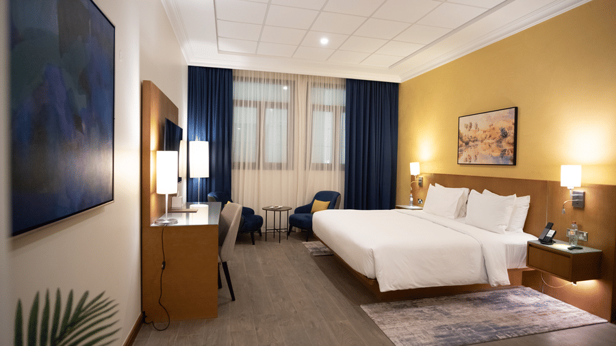 Spacious bedroom with large bed with white linens, two stools, yellow feature wall, and windows with blue and sheer curtains.