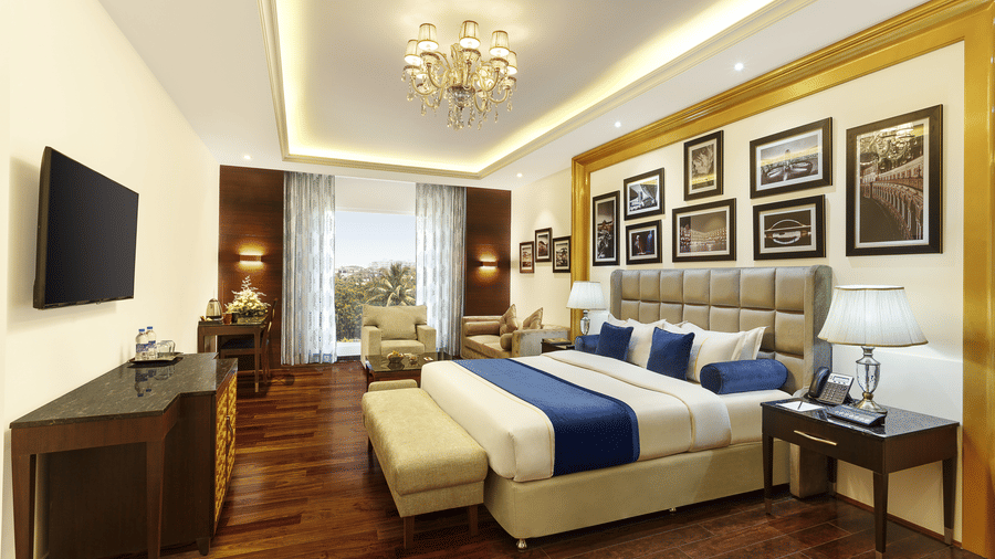 The executive suite bedroom with a king size bed, seating area, television, wooden flooring, framed wall decor and elegant lighting at La Marvella, Bengaluru.