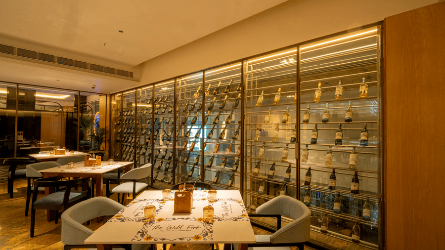 Dining table and chairs at The Wild Fork restaurant in La Marvella, Bengaluru which also featurings a glass shelve where several liquor bottles are placed.