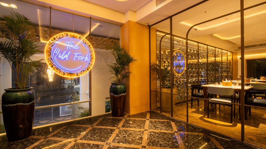 Entrance of The Wild Fork at La Marvella, Bengaluru featuring the logo of the restaurant with two flower pots on either side. It also features glass doors leading to the dining space.