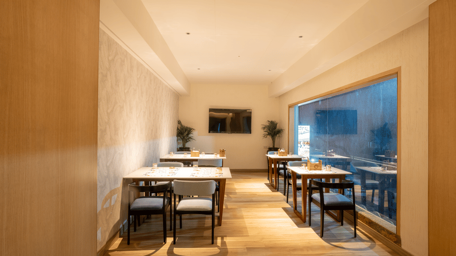 A section of a dining area at La Marvella, Bengaluru featuring table and chairs placed on either side and there is a flat TV screen at the end.