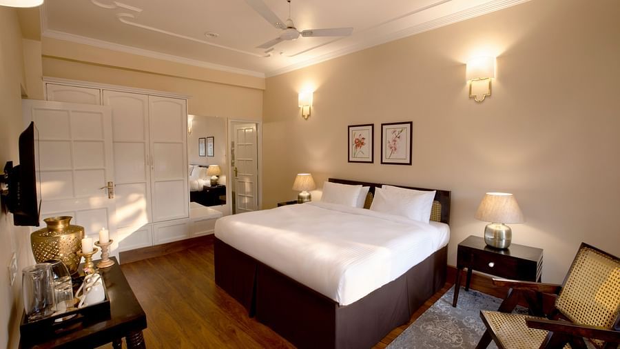 An image of super deluxe room featuring a king size bed, bedside table on either side, a flat TV screen, a table where antique items are placed, and  a chair in one corner of the room- Lamrin Boutique Cottages Rishikesh
