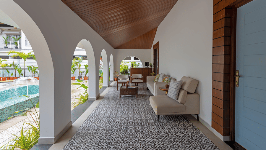 An indoor lounge area with a tiled black-and-white patterned floor, white seating, and arched doorways leading to an outdoor pool and garden area - Lamrin Morjim Goa