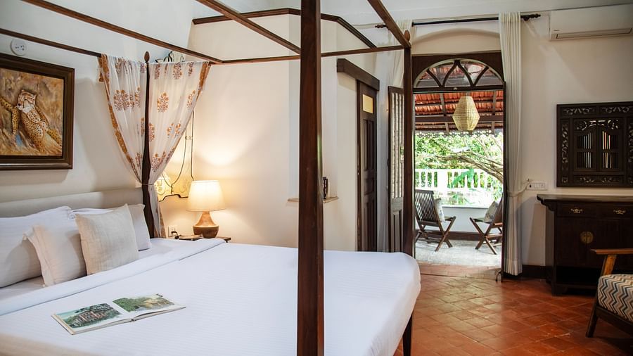 An image of a bed with four posters, a bedside table with lamp, a wardrobe, and open wooden door at Lamrin Ucassaim Hotel, Goa