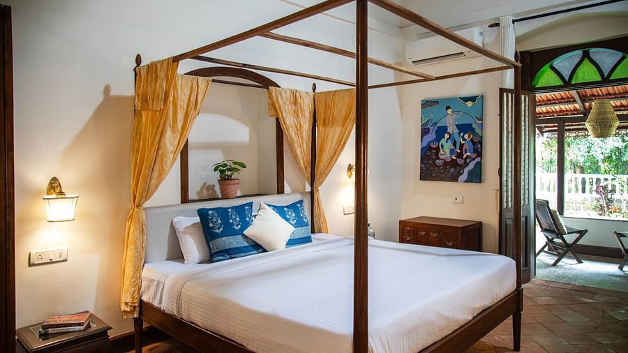An image of four poster bed with yellow curtains,bedside table with a lamp, a cabinet at one corner, and an attached balcony where a chair is place - Lamrin Ucassaim Hotel, Goa