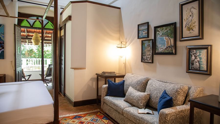 An image of a beautiful grand bedroom  with seating area, photo frames hanging on the wall and there is also a table with lamp - Lamrin Ucassaim Hotel, Goa