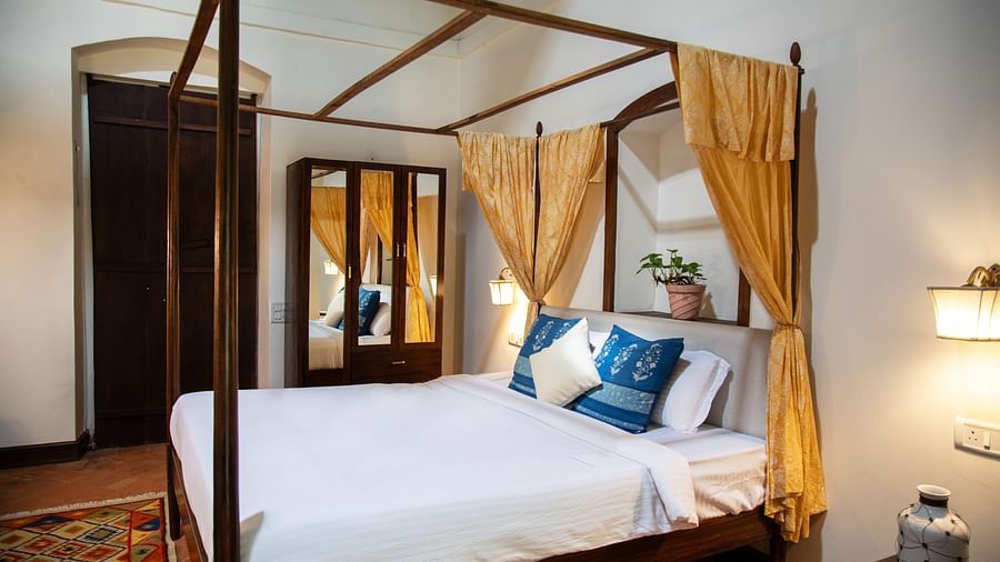 An image of a four poster bed, bedside table, wooden dressing table, and other wooden furnishings at Lamrin Ucassaim Hotel, Goa