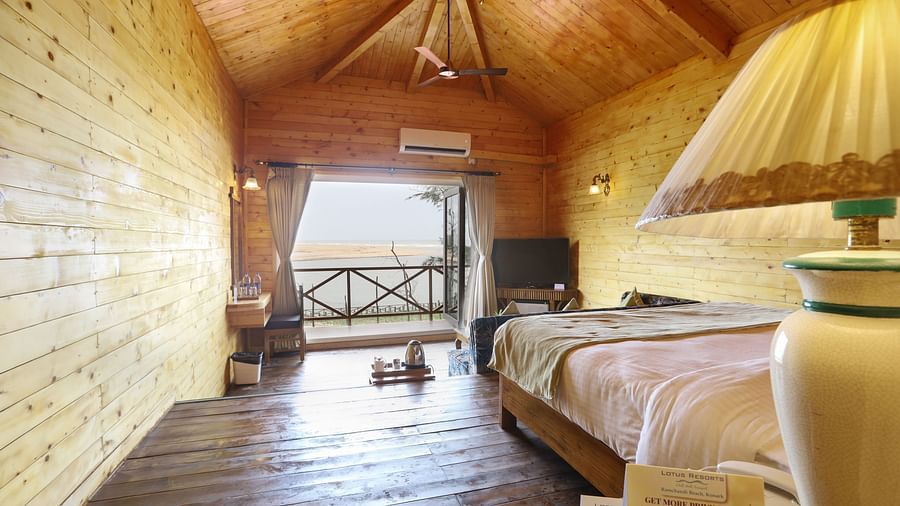A bright and airy room at Lotus Eco Beach Resort Konark, featuring wooden walls, a cosy bed, and doors opening to a balcony with a view, all under a high ceiling with a fan.