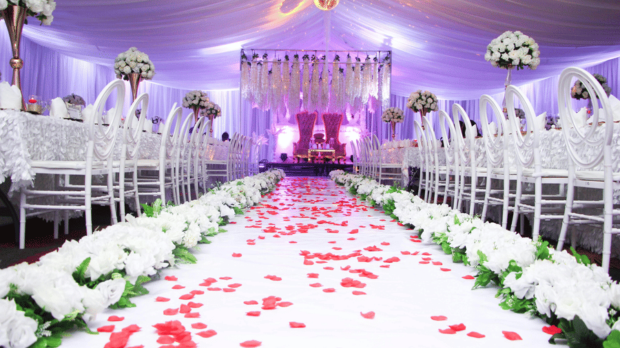 A white carpet aisle covered in red rose petals with white chairs on both sides and floral arrangements on stands leading toward a decorated stage.