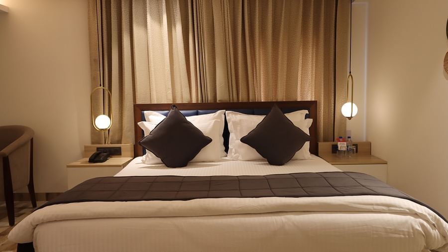 Front view of the plush bed in Deluxe King with two bedside tables and illuminating lamp, partial view of the cabinet - Mastiff Hotel, Dhanbad 