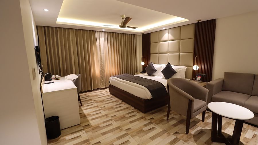 Side angle view of a cozy bed with bed side table and lamp, small desk for storage and  TV, sofa set with a coffee table for Studio room - Mastiff Hotel, Dhanbad