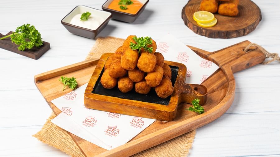 A wooden tray holds a metal basket of round fried items, surrounded by small dipping sauce bowls, a lemon wedge, and a few more pieces.