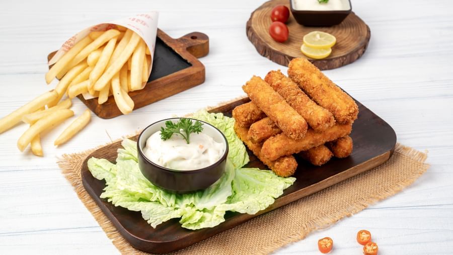 A wooden plate holds a serving of french fries, fried cheese sticks, and a bowl of dipping sauce, with bread sticks on the side.