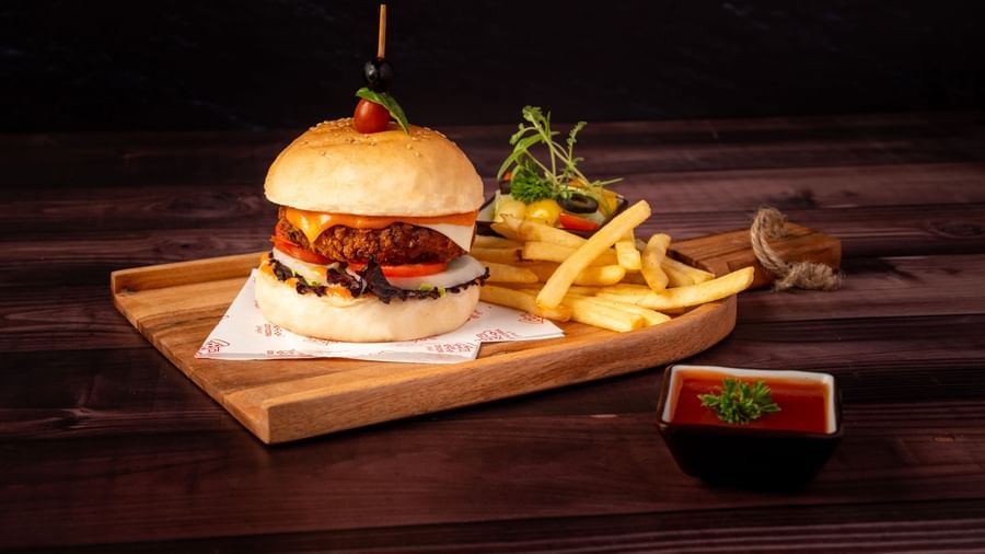 A large burger with a stick through it is on a wooden tray with a side of french fries and a small dish of dipping sauce.