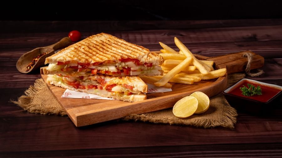 A grilled sandwich, cut in half, is served with french fries, a lemon wedge, and a small bowl of sauce on a wooden board.