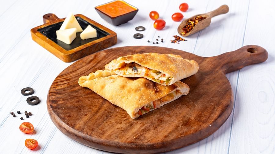 A wooden board holds two halves of a calzone, surrounded by cheese wedges, tomatoes, olives, and a bowl of sauce.