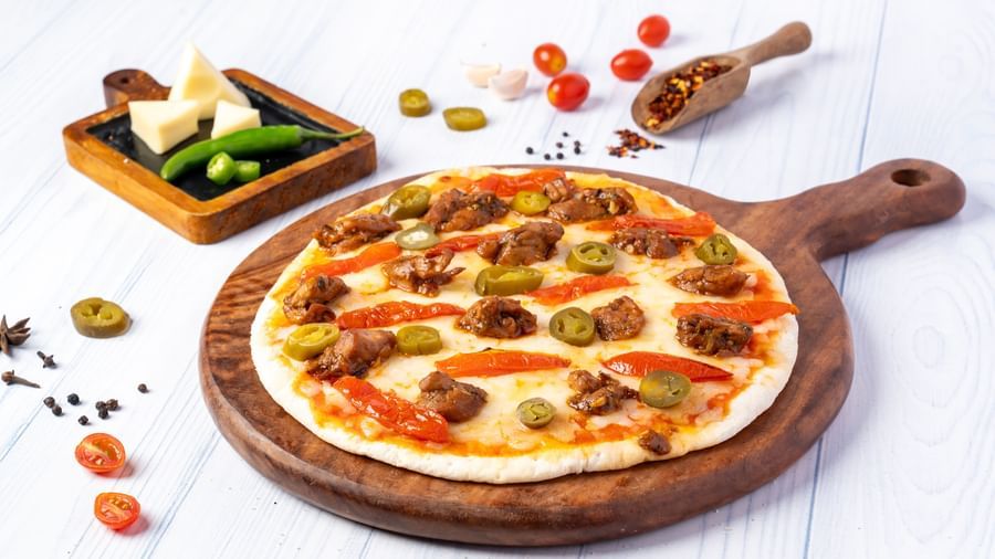 A round pizza topped with meat, cheese, and sliced peppers sits on a wooden board, with cheese, chili flakes, and cherry tomatoes nearby.