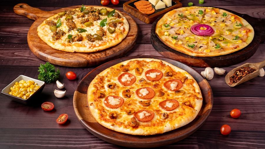 Three different round pizzas are displayed on a wooden surface, surrounded by ingredients like corn, cherry tomatoes, and cheese.