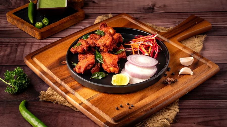 A plate on a wooden board holds fried chicken pieces with a side of sliced onions, a lime wedge, and shredded vegetables.
