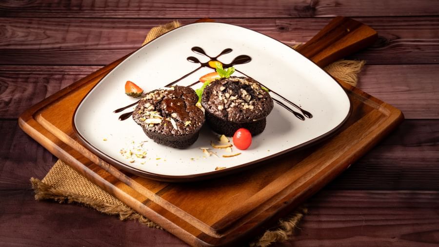 A triangular plate on a wooden board holds two chocolate muffins with nuts, drizzled with chocolate sauce and garnished with strawberries.